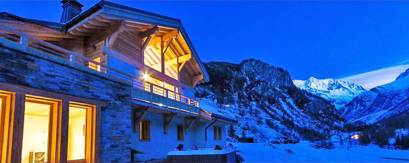 Chalet Cairn im Schnee zur blauen Stunde, erleuchtete Fenster, Menschen im Außen-Jacuzzi vor dem Haus