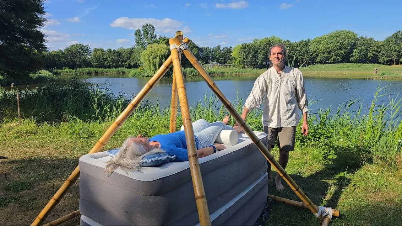 Gerald steht am Bambus-Dreibein neben einer Person auf dem Luftkissen, dahinter ein Teich